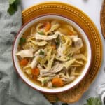 Creamy chicken noodle soup with vegetables in a white bowl, garnished with herbs on a rustic table setting.