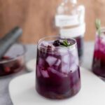 Fresh berry-infused purple cocktail with ice cubes and a sprig of rosemary, served in a clear glass. Perfect for summer drinks and berry recipes.