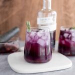 Fresh purple berry iced beverage with ice cubes and a sprig of rosemary, served in a clear glass, with a bottle of gin and additional drinks in the background, on a gray surface.