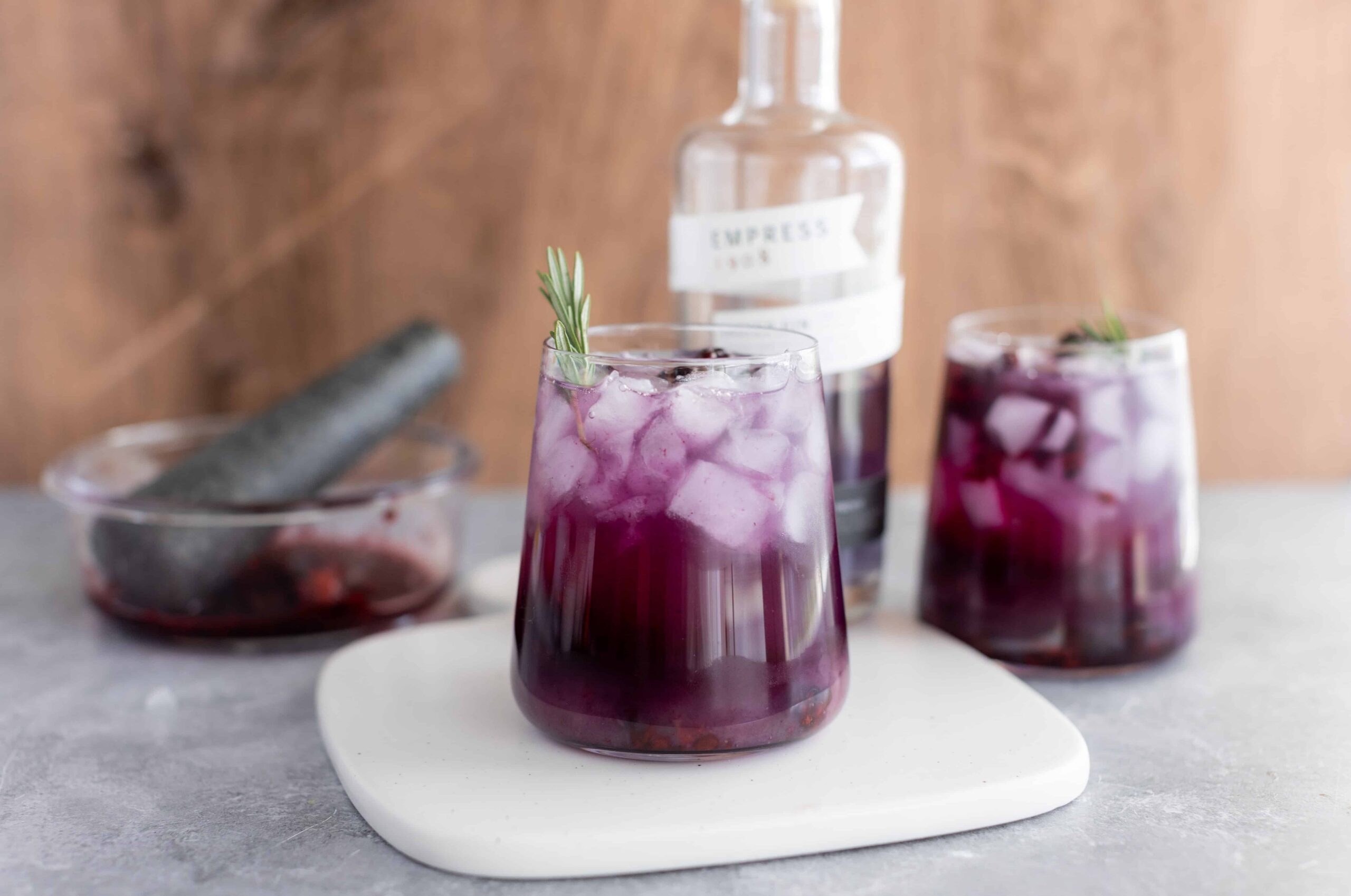 Fresh purple berry iced beverage with ice cubes and a sprig of rosemary, served in a clear glass, with a bottle of gin and additional drinks in the background, on a gray surface.