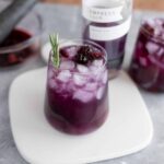 Fresh berry infusions with ice in a tall glass, garnished with rosemary, featuring ingredients like mixed berries and herbs for homemade summer drinks.