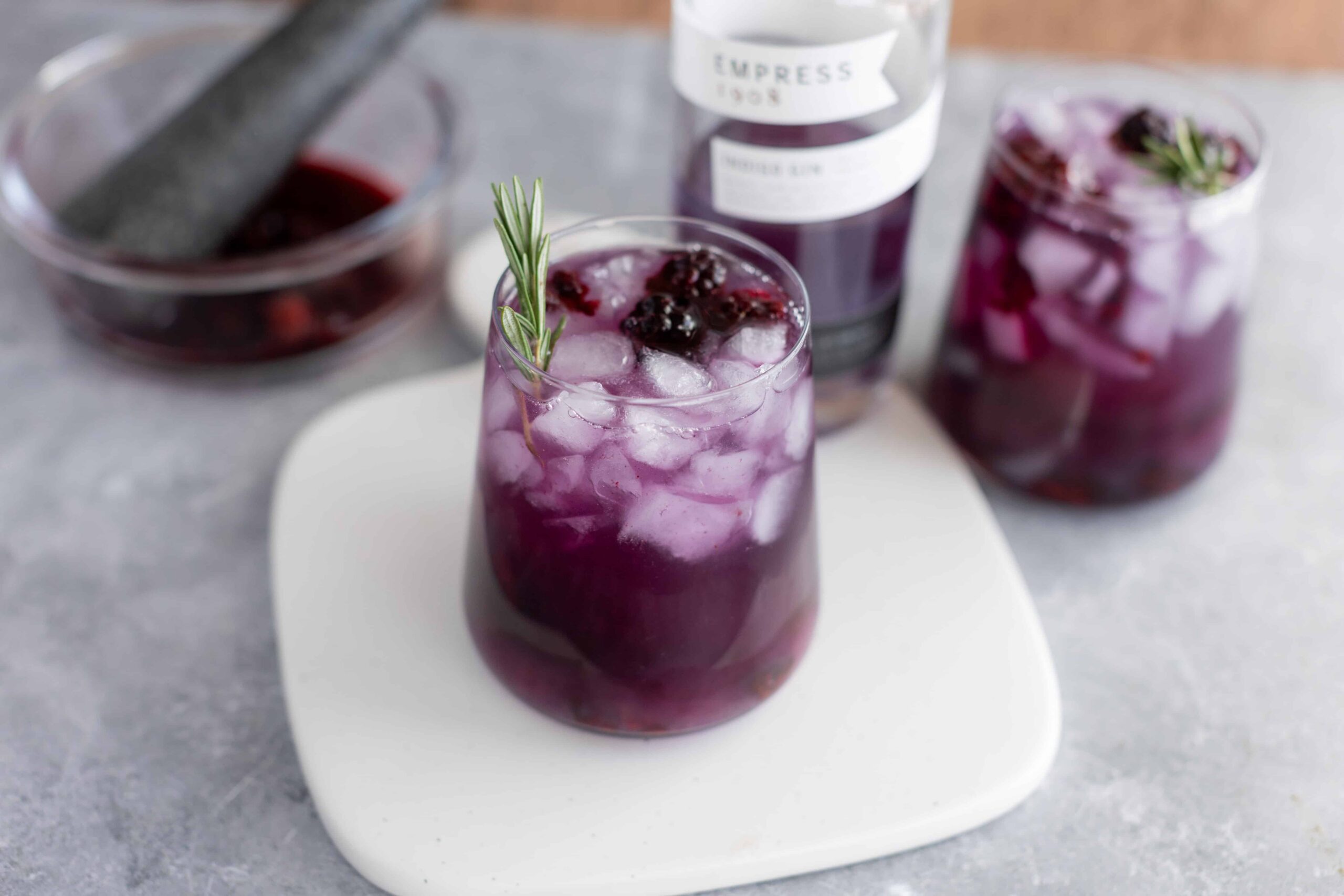 Fresh berry infusions with ice in a tall glass, garnished with rosemary, featuring ingredients like mixed berries and herbs for homemade summer drinks.
