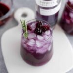 Refreshing blackberry lavender mocktail with ice cubes, garnished with a sprig of fresh rosemary, served in a clear glass on a light gray surface.