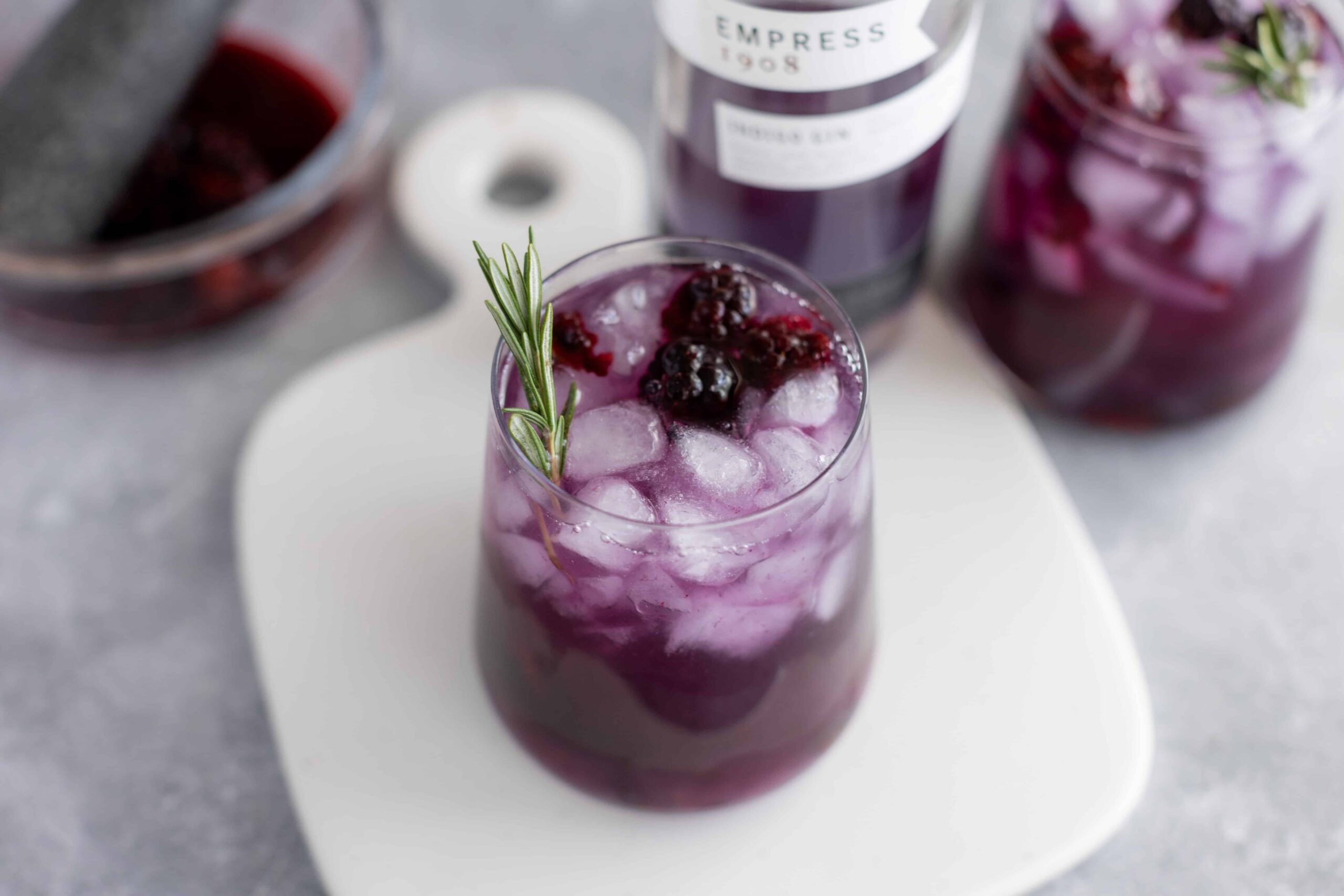 Refreshing blackberry lavender mocktail with ice cubes, garnished with a sprig of fresh rosemary, served in a clear glass on a light gray surface.