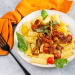 Creamy penne pasta with cherry tomatoes, fresh basil, and roasted vegetables on a white plate, served with a black fork and a burnt orange napkin on a light gray background.