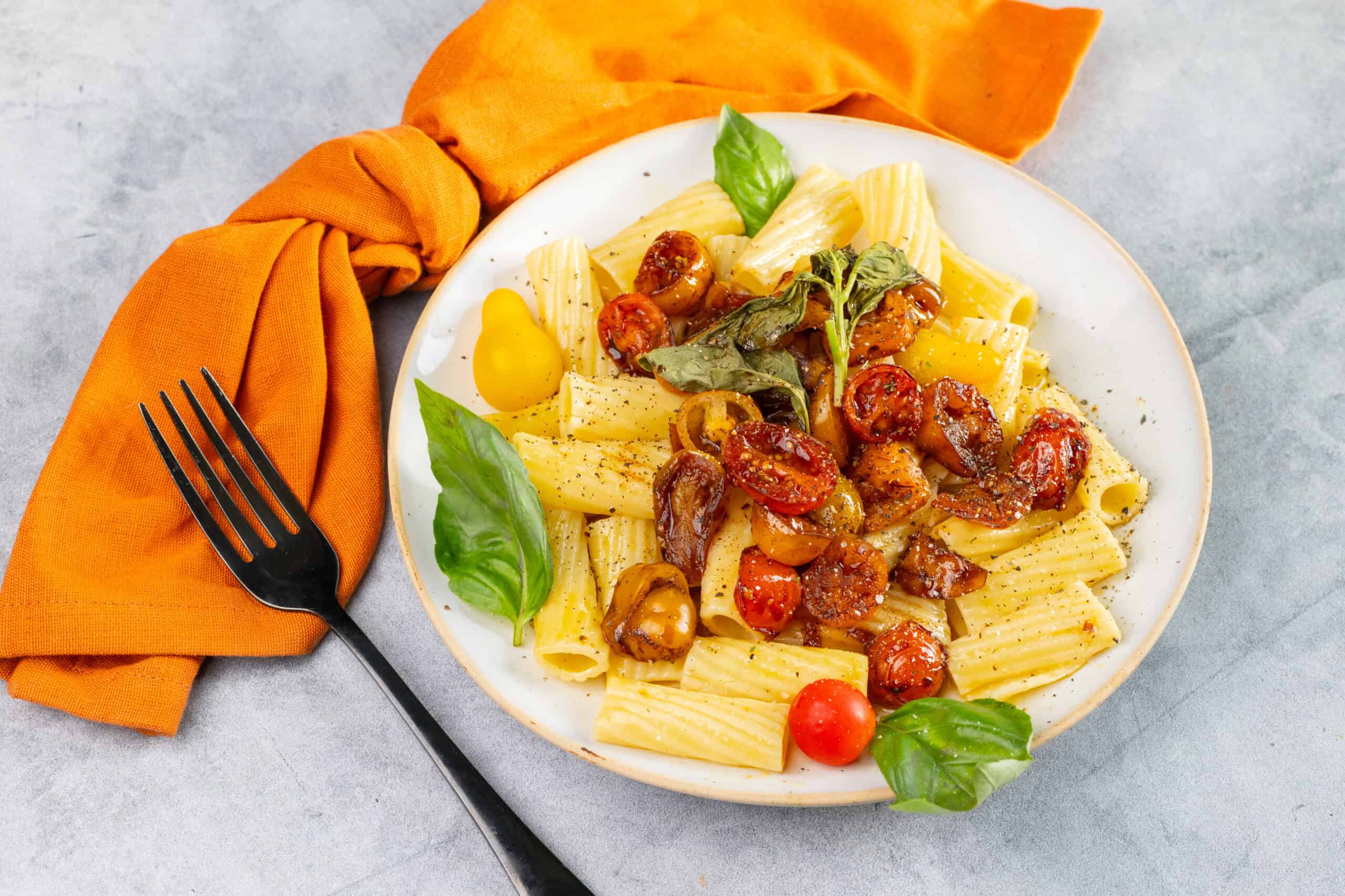 Creamy penne pasta with cherry tomatoes, fresh basil, and roasted vegetables on a white plate, served with a black fork and a burnt orange napkin on a light gray background.