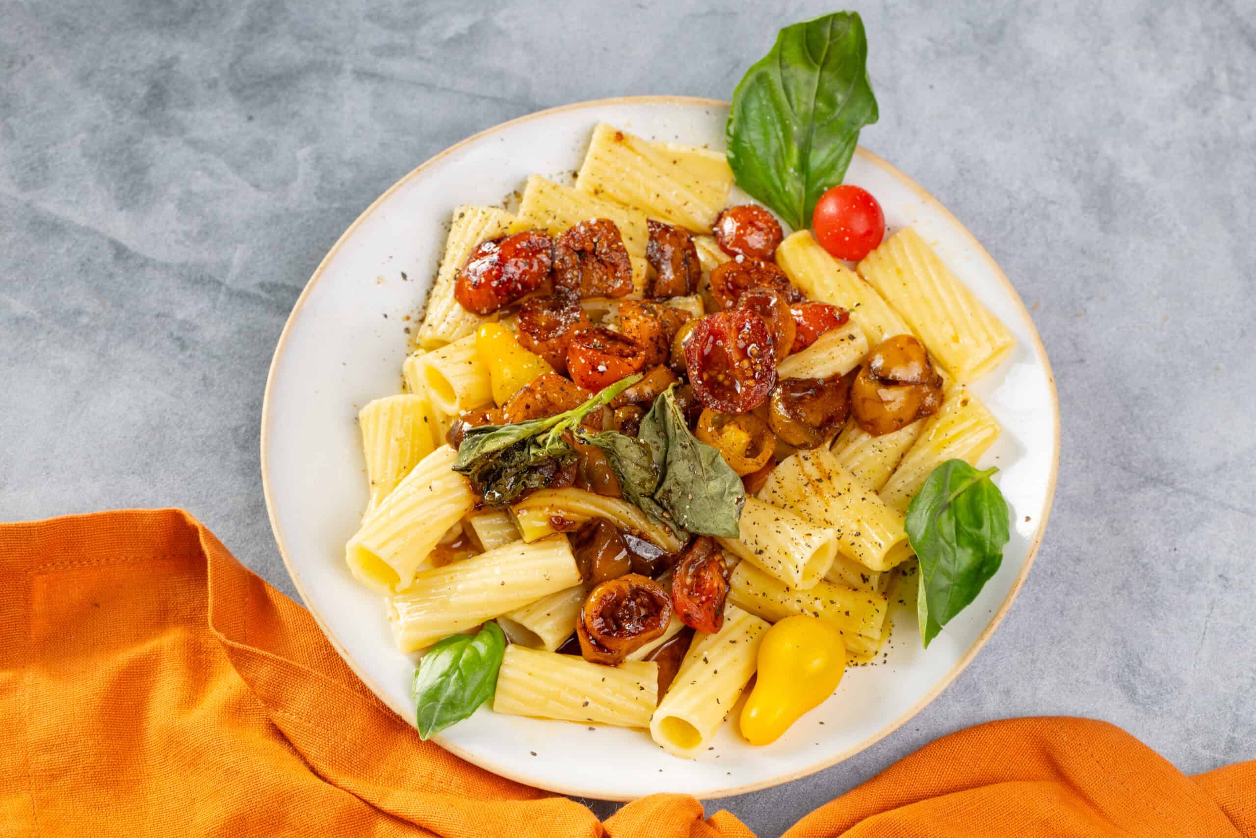 Roasted cherry tomato pasta with fresh basil and mushrooms on white plate, served with colorful cherry tomatoes and basil leaves for authentic Italian flavor.