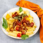 Roasted cherry tomato pasta with fresh basil leaves on a white plate, served with a vibrant orange cloth, garnished with herbs and black pepper, perfect for a delicious Italian dinner.