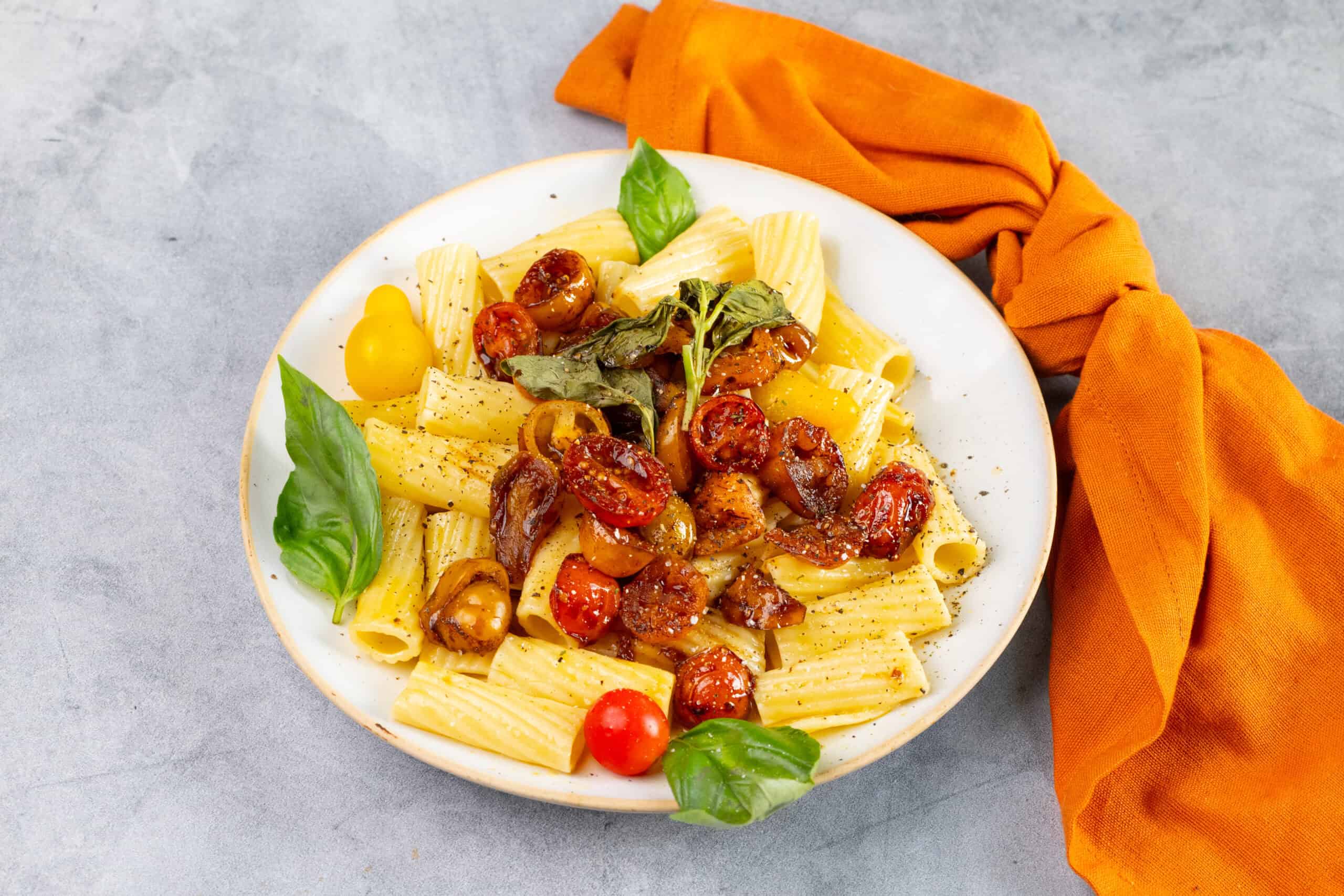 Roasted cherry tomato pasta with fresh basil leaves on a white plate, served with a vibrant orange cloth, garnished with herbs and black pepper, perfect for a delicious Italian dinner.