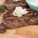 Juicy grilled steak topped with creamy blue cheese butter, served on a wooden cutting board with herbs and cheese in the background for a delicious recipe presentation.