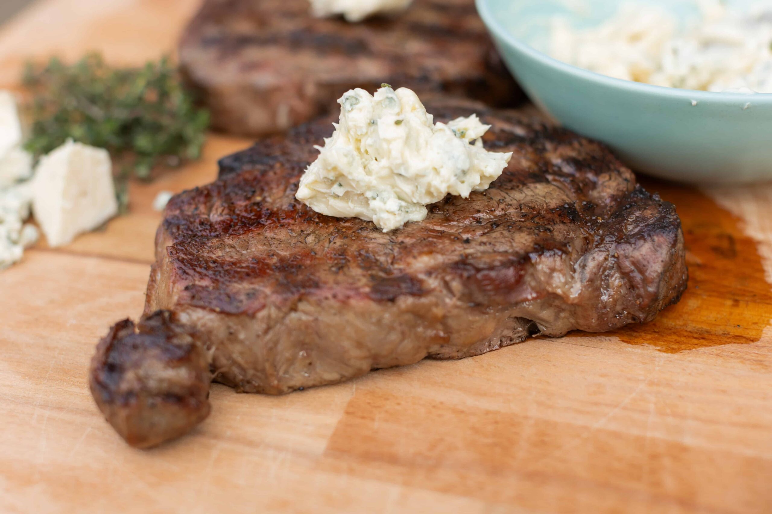 Juicy grilled steak topped with creamy blue cheese butter, served on a wooden cutting board with herbs and cheese in the background for a delicious recipe presentation.