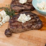 Juicy grilled steak topped with blue cheese butter on a wooden cutting board, with additional cheese, herbs, and a bowl of blue cheese crumbles, showcasing a delicious steak dinner.