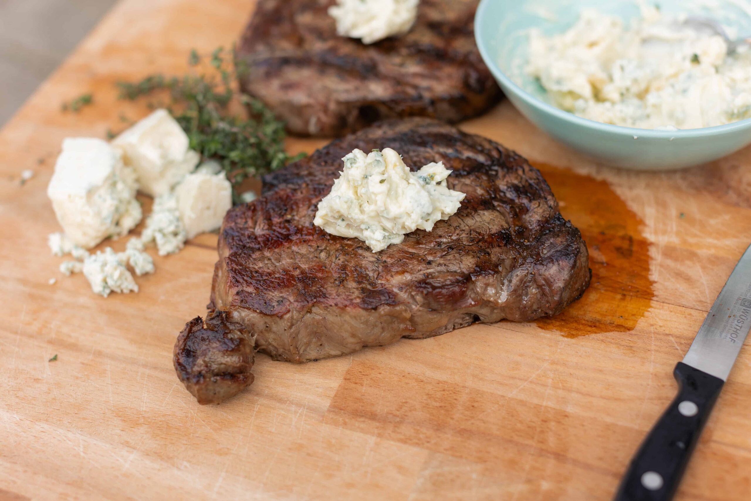 Juicy grilled steak topped with blue cheese butter on a wooden cutting board, with additional cheese, herbs, and a bowl of blue cheese crumbles, showcasing a delicious steak dinner.