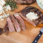 Juicy grilled steak topped with herb butter, sliced on a wooden cutting board with blue cheese crumbles, fresh herbs, and a carving knife, perfect for a flavorful meat dinner.