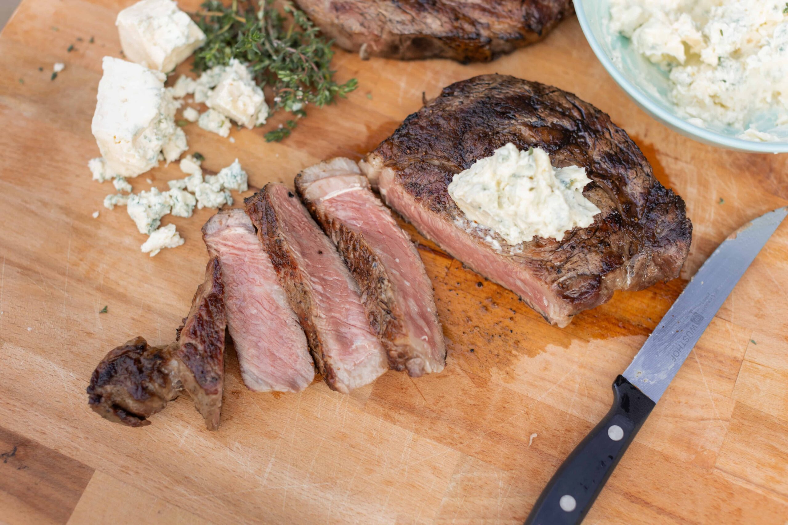 Juicy grilled steak topped with herb butter, sliced on a wooden cutting board with blue cheese crumbles, fresh herbs, and a carving knife, perfect for a flavorful meat dinner.