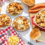 Creamy baked macaroni and cheese with ground beef, vegetables, and toasted bread crumbs served in a red casserole dish with bread rolls and fresh carrots on a checkered tablecloth.