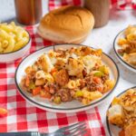 Savory casserole with ground beef, pasta, cheese, and vegetables on a white plate with additional servings and bread on a red checkered tablecloth, comfort food meal for dinner or family gathering.