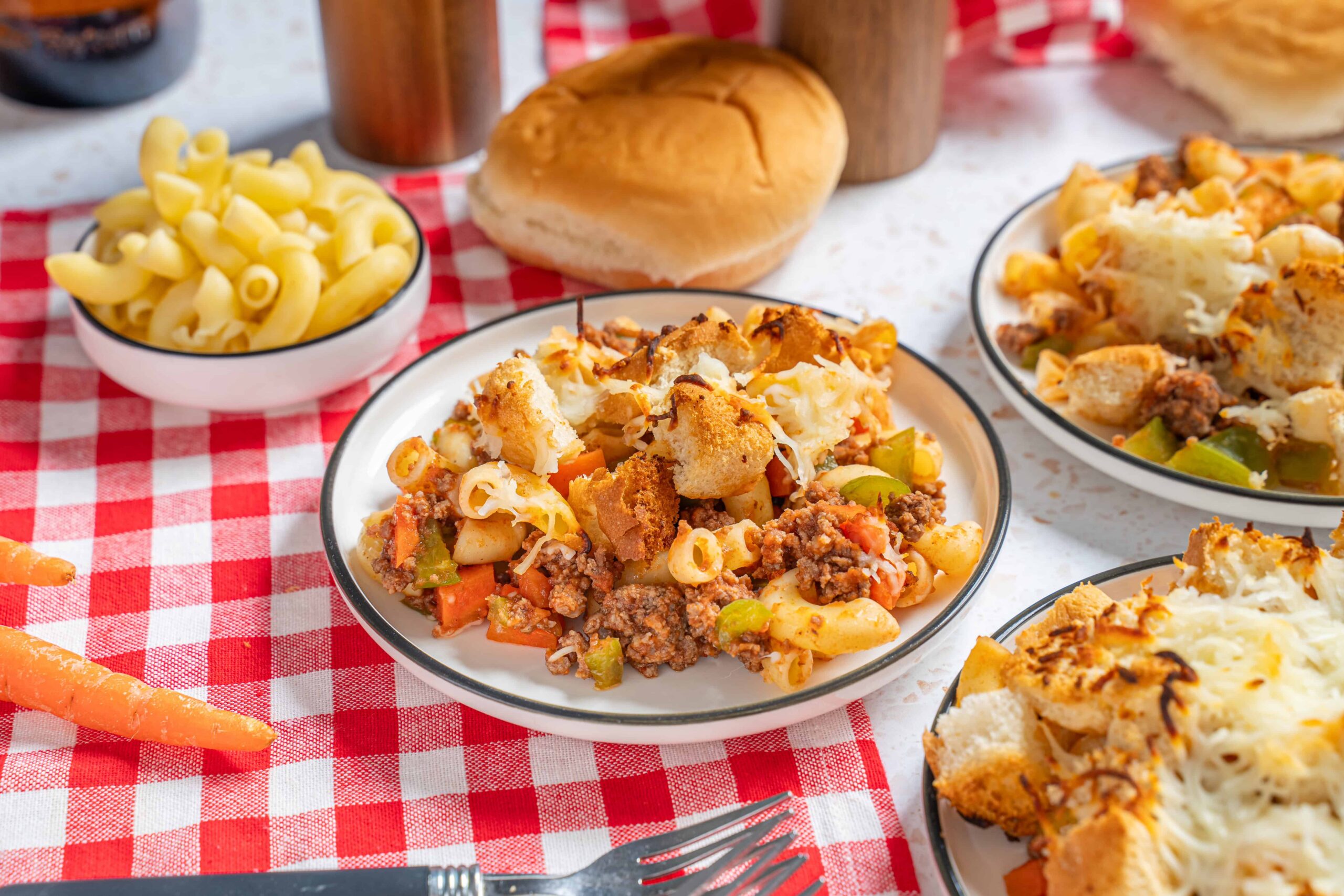 Savory casserole with ground beef, pasta, cheese, and vegetables on a white plate with additional servings and bread on a red checkered tablecloth, comfort food meal for dinner or family gathering.