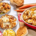 Creamy hamburger casserole with cheese, ground beef, pasta, and fresh bread on a wooden board. Perfect comfort food for lunch or dinner with a side of carrots and baked bread rolls.