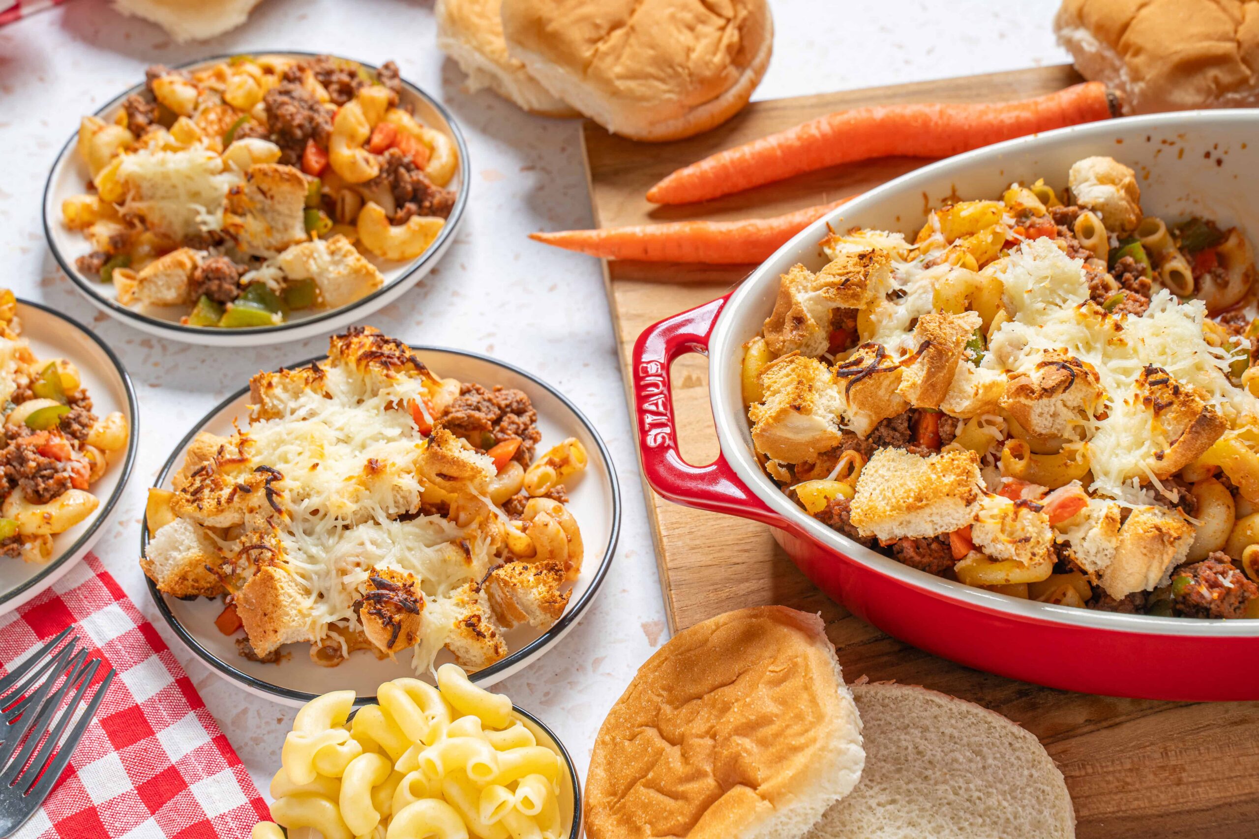 Creamy hamburger casserole with cheese, ground beef, pasta, and fresh bread on a wooden board. Perfect comfort food for lunch or dinner with a side of carrots and baked bread rolls.