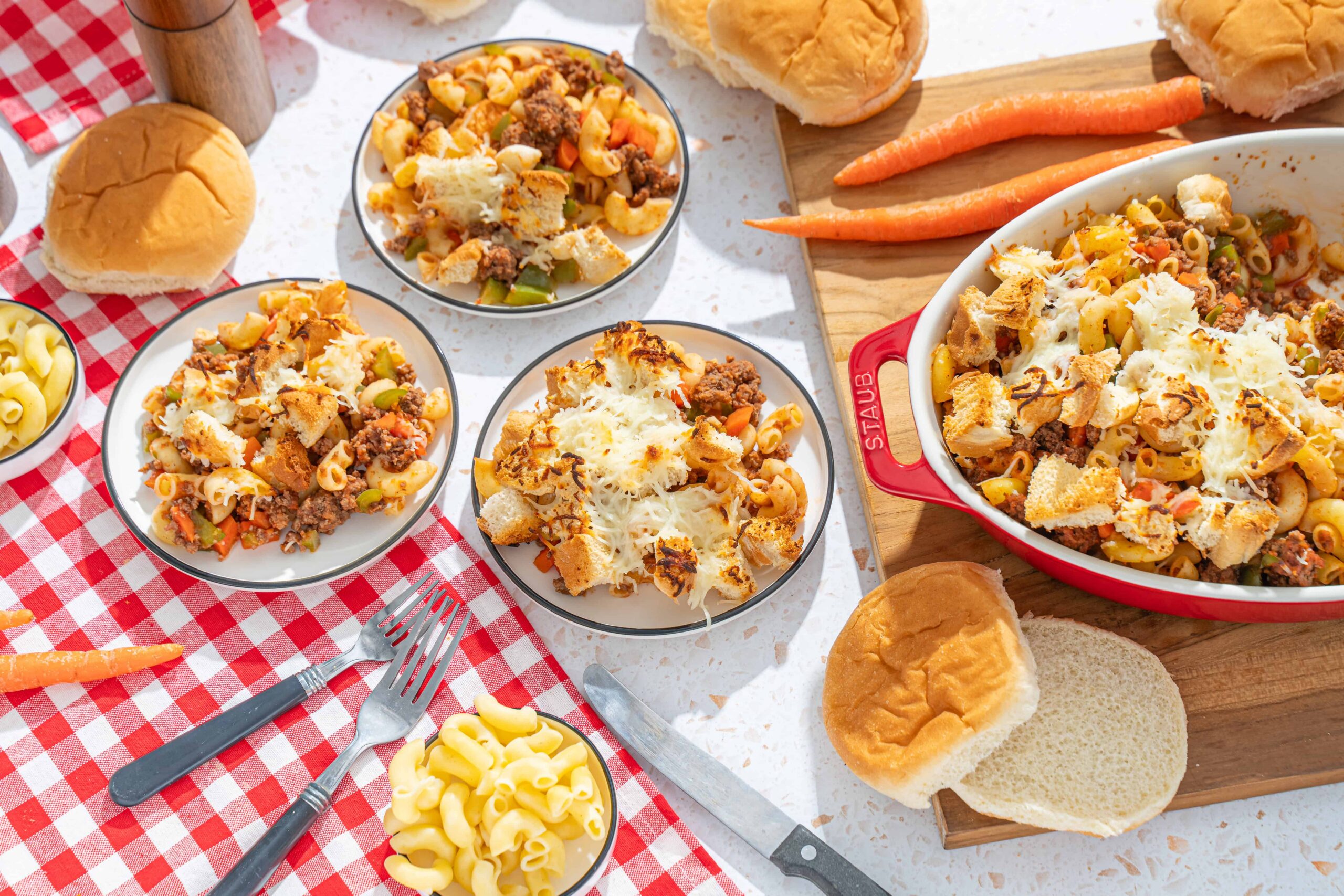 Creamy baked macaroni and cheese with ground beef, vegetables, and toasted bread crumbs served in a red casserole dish with bread rolls and fresh carrots on a checkered tablecloth.