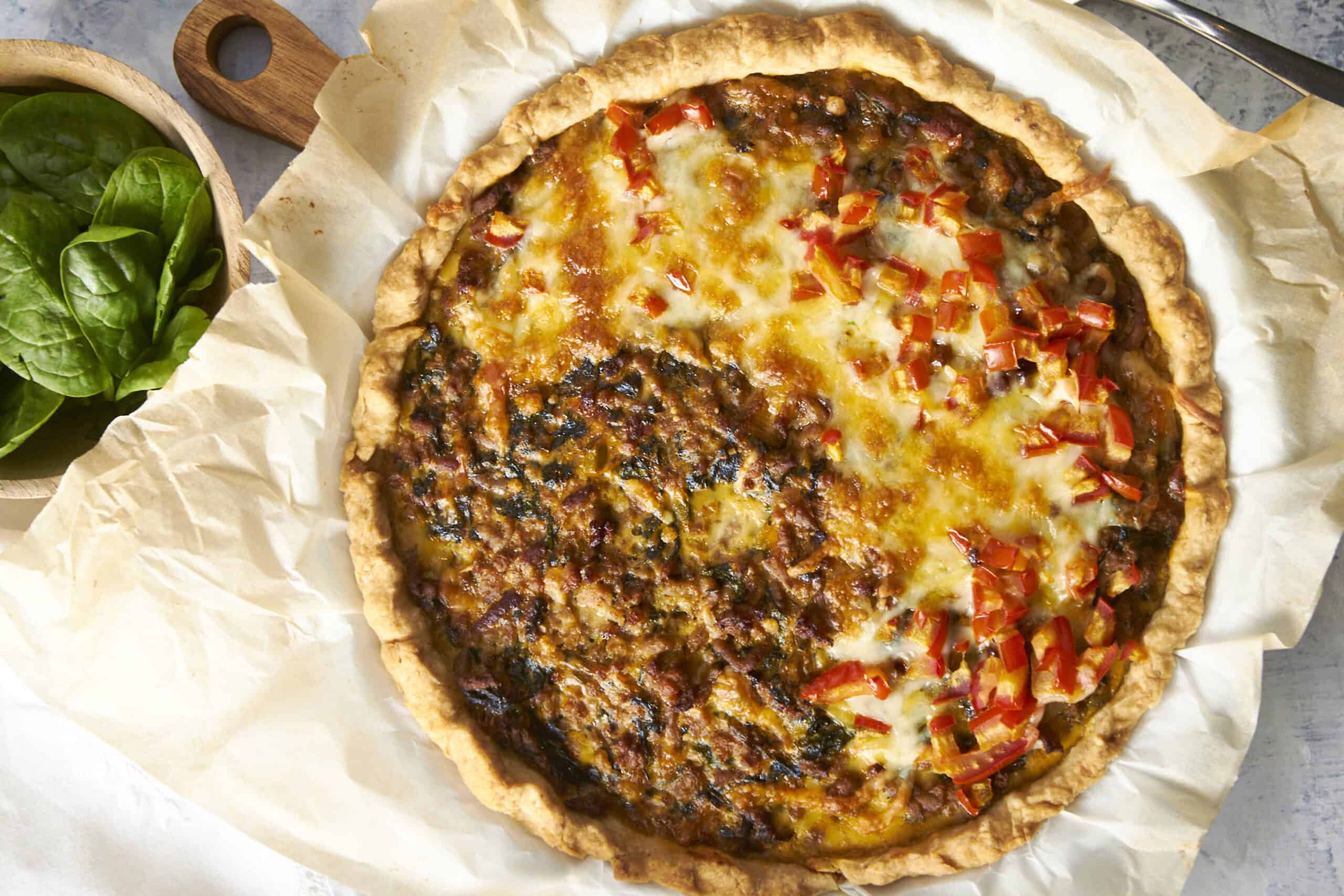 Golden-brown homemade savory beef and vegetable pie with melted cheese and fresh diced red peppers on top, served on parchment paper with a side of fresh basil leaves, perfect for a comforting meal.