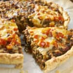 Golden-brown savory meat and cheese stuffed savory pie with a flaky crust, topped with diced red peppers, on parchment paper.