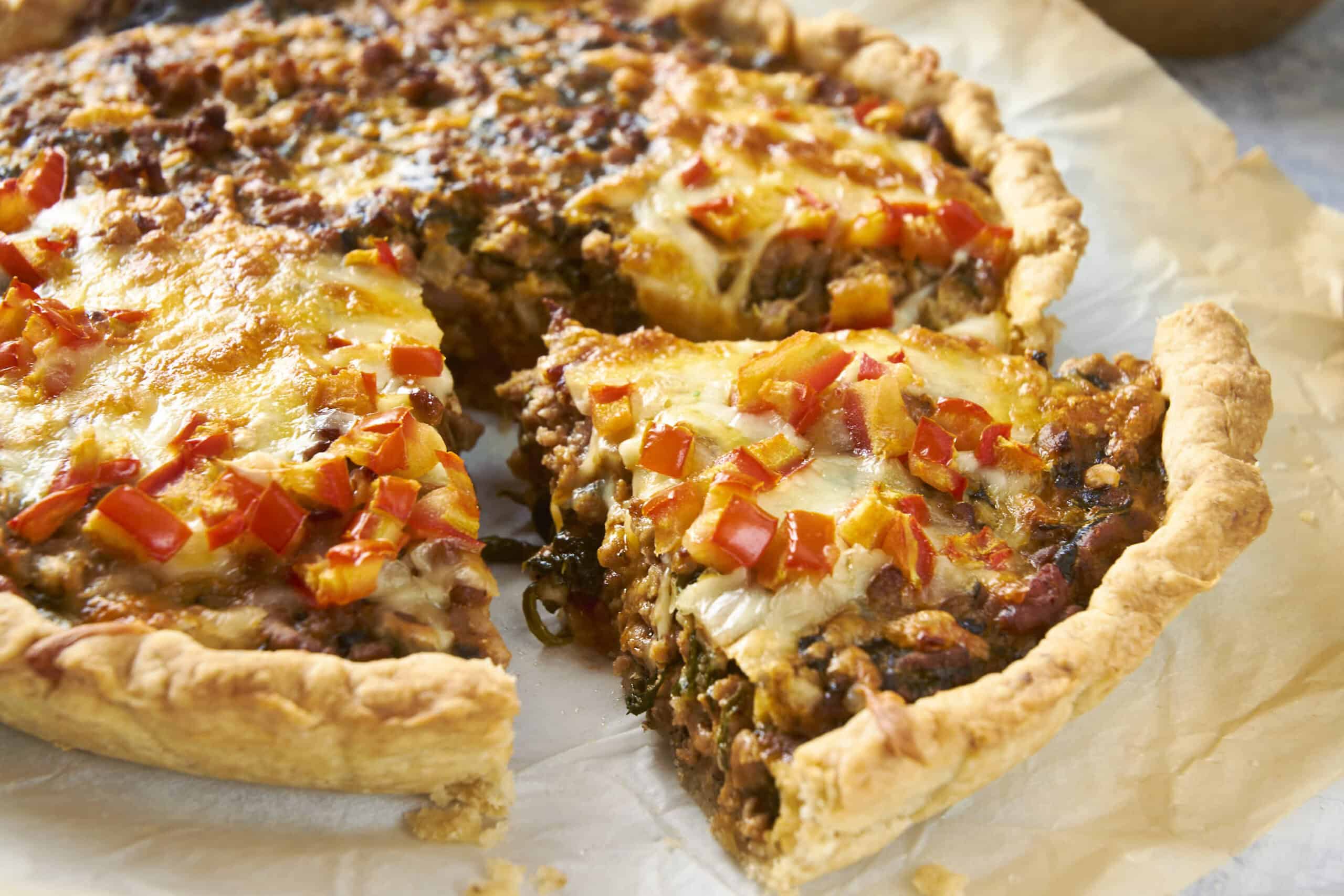 Golden-brown savory meat and cheese stuffed savory pie with a flaky crust, topped with diced red peppers, on parchment paper.