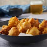 Crispy golden battered fried chicken pieces served in a black bowl on a textured surface, with a side of beer and dipping sauce in the background, perfect for comfort food and snack recipes.