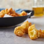 Crispy fried cauliflower florets on a textured surface with a bowl of fried chicken in the background and a glass of beer, perfect for snack or appetizer, showcasing delicious comfort food.