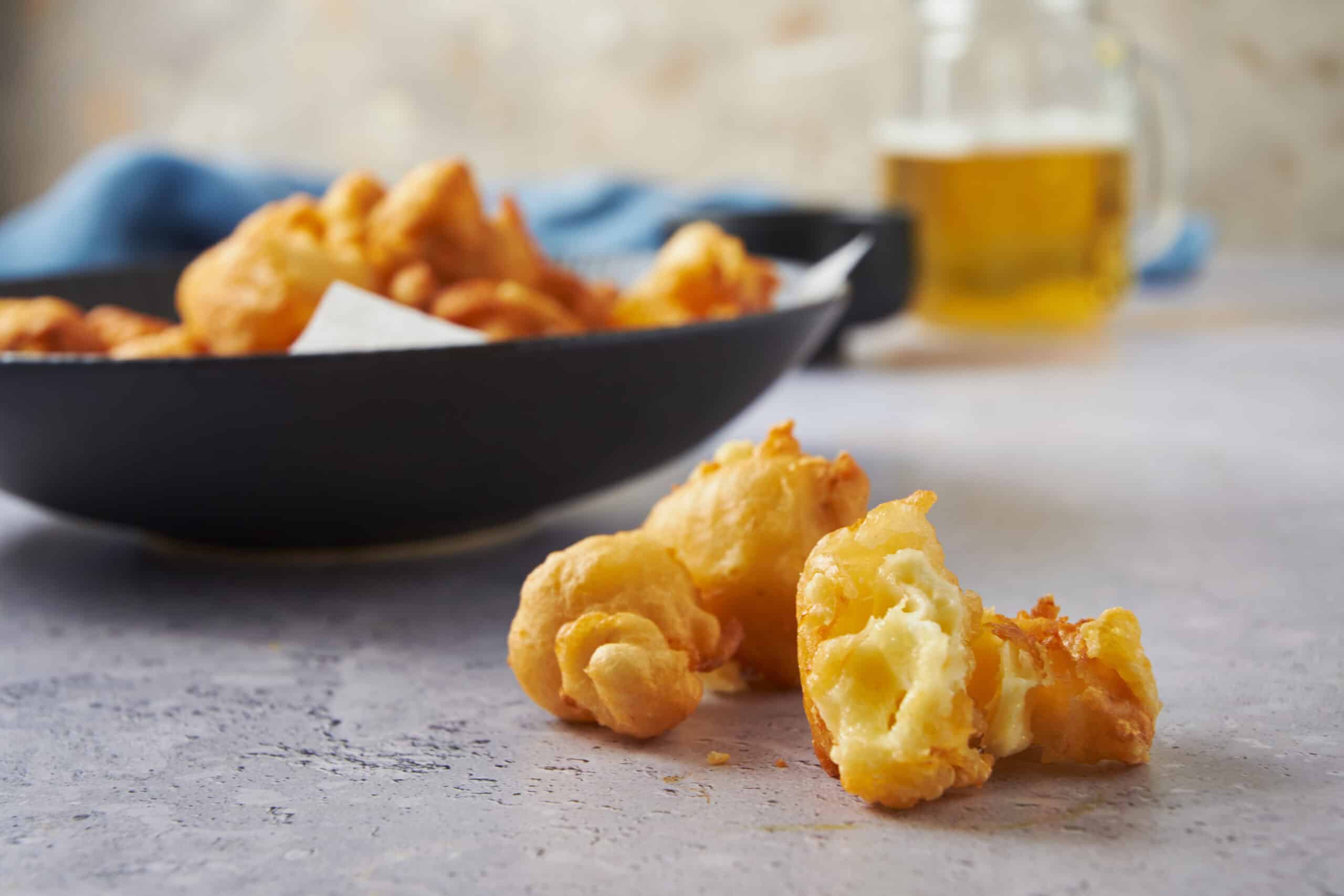 Crispy fried cauliflower florets on a textured surface with a bowl of fried chicken in the background and a glass of beer, perfect for snack or appetizer, showcasing delicious comfort food.