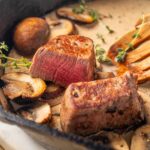 Juicy pan-seared steak with mushrooms in a creamy sauce, garnished with fresh thyme, served with a wooden spatula, close-up image of a delicious beef filet for recipe and cooking ideas.