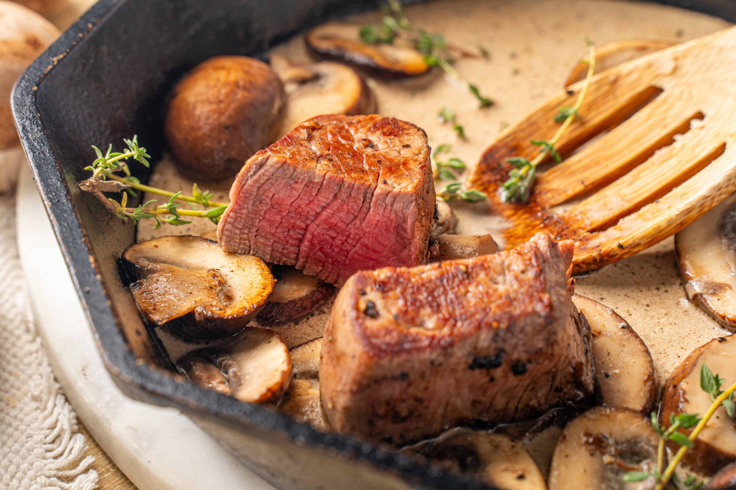 Juicy pan-seared steak with mushrooms in a creamy sauce, garnished with fresh thyme, served with a wooden spatula, close-up image of a delicious beef filet for recipe and cooking ideas.