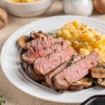 Juicy medium-rare steak with sautéed mushrooms and creamy mashed potatoes garnished with fresh herbs, served on a white plate for a delicious and hearty meal.