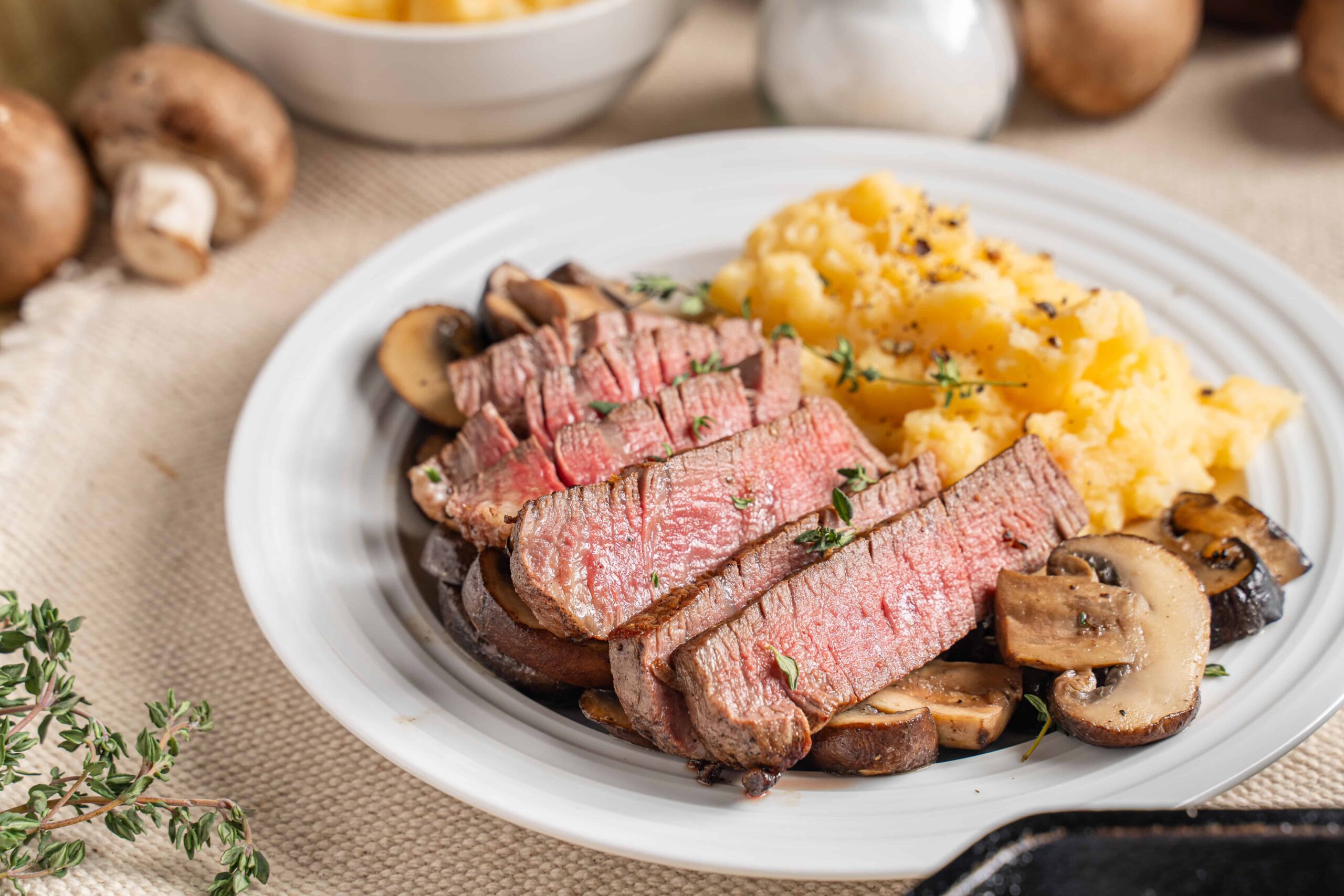 Juicy medium-rare steak with sautéed mushrooms and creamy mashed potatoes garnished with fresh herbs, served on a white plate for a delicious and hearty meal.