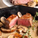 Juicy medium-rare steak with sautéed mushrooms in a creamy sauce, garnished with fresh thyme in a cast iron skillet for a delicious gourmet meal.