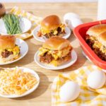 Savory breakfast casserole with eggs, sausage, cheese, and bread baked in a red dish, served with fresh eggs, shredded cheese, and chives on a wooden table. Perfect for brunch or quick family breakfast.