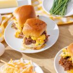 Savory breakfast sandwiches with scrambled eggs, cooked sausage, and cheese in soft, toasted buns on white plates, surrounded by eggs, shredded cheese, and fresh chives.