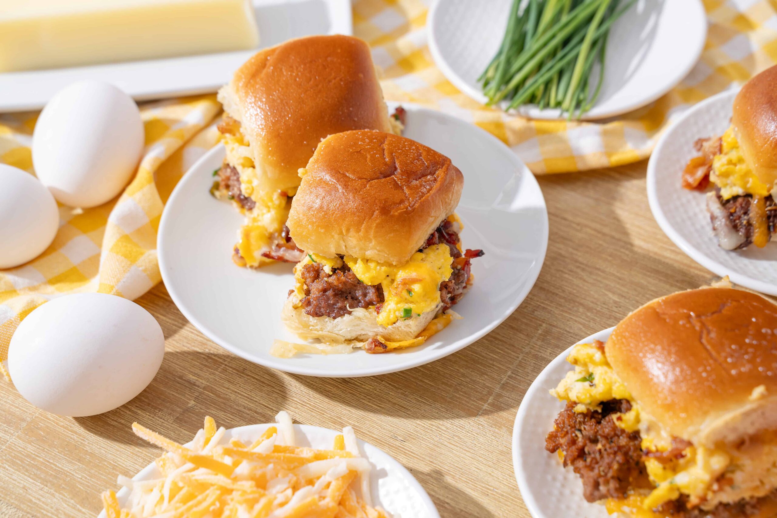 Savory breakfast sandwiches with scrambled eggs, cooked sausage, and cheese in soft, toasted buns on white plates, surrounded by eggs, shredded cheese, and fresh chives.