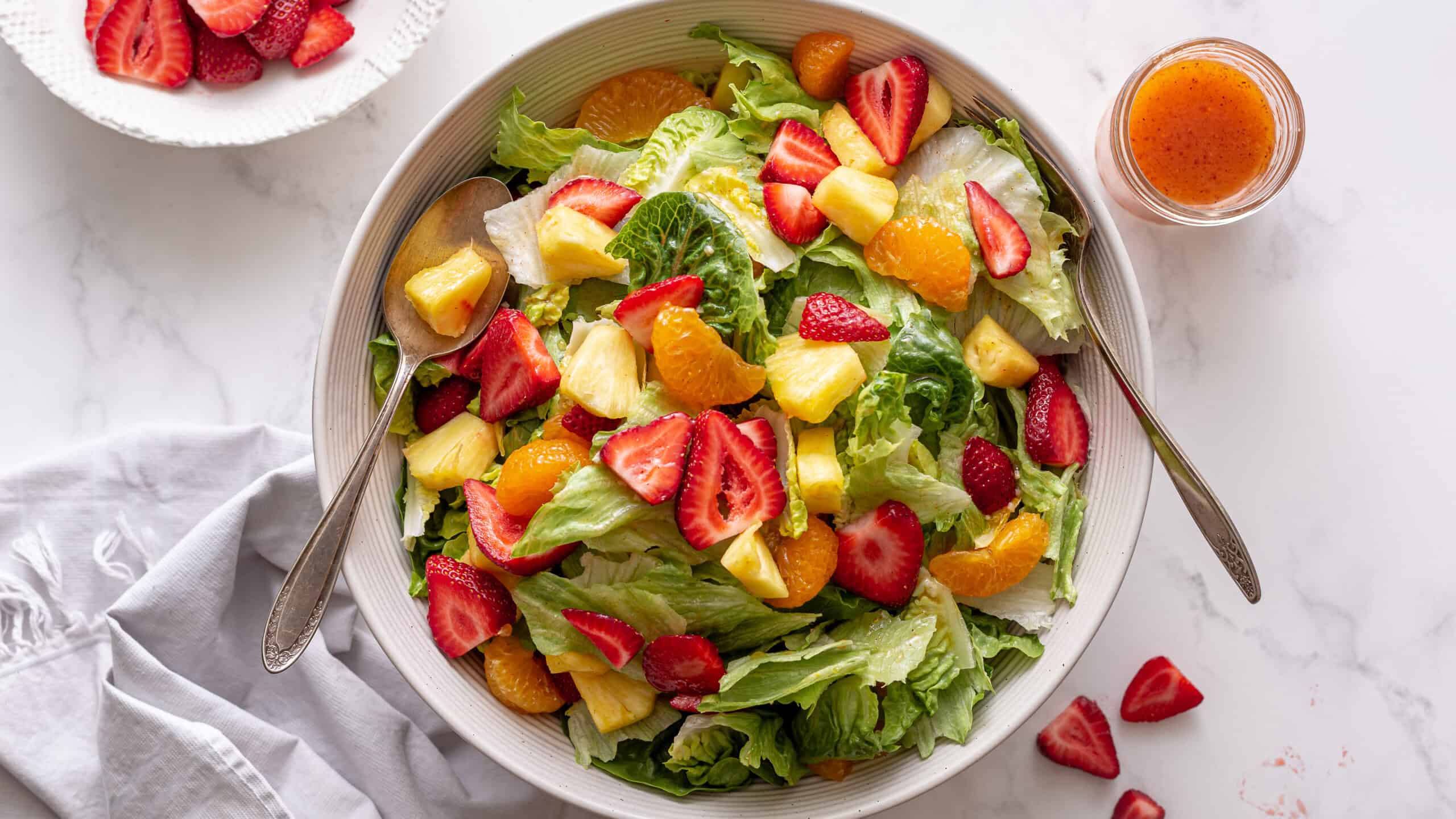 Fresh mixed fruit and vegetable salad with strawberries, pineapple, oranges, and lettuce on a white plate with a side of vinaigrette dressing.
