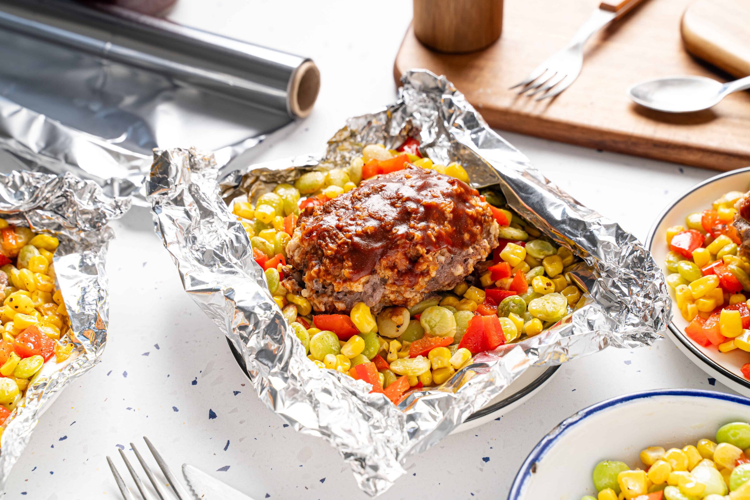 Juicy meatloaf with vegetables wrapped in aluminum foil, ready to bake, on a white kitchen countertop with cooking tools and dishes nearby.