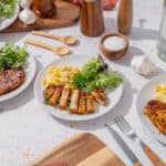 Grilled chicken with fresh salad, scrambled eggs, and herbs on white plates served at a bright tabletop with cooking ingredients and utensils.