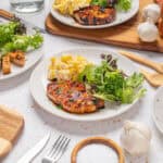 Grilled salmon with fresh greens and mashed potatoes on white plates at a rustic table, featuring garlic, salt, and wooden serving utensils, showcasing healthy seafood dinner options.