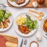 Grilled chicken breast with fresh side salad, corn salad, and mashed potatoes on white plates, served on a white textured surface for healthy dinner or lunch ideas.