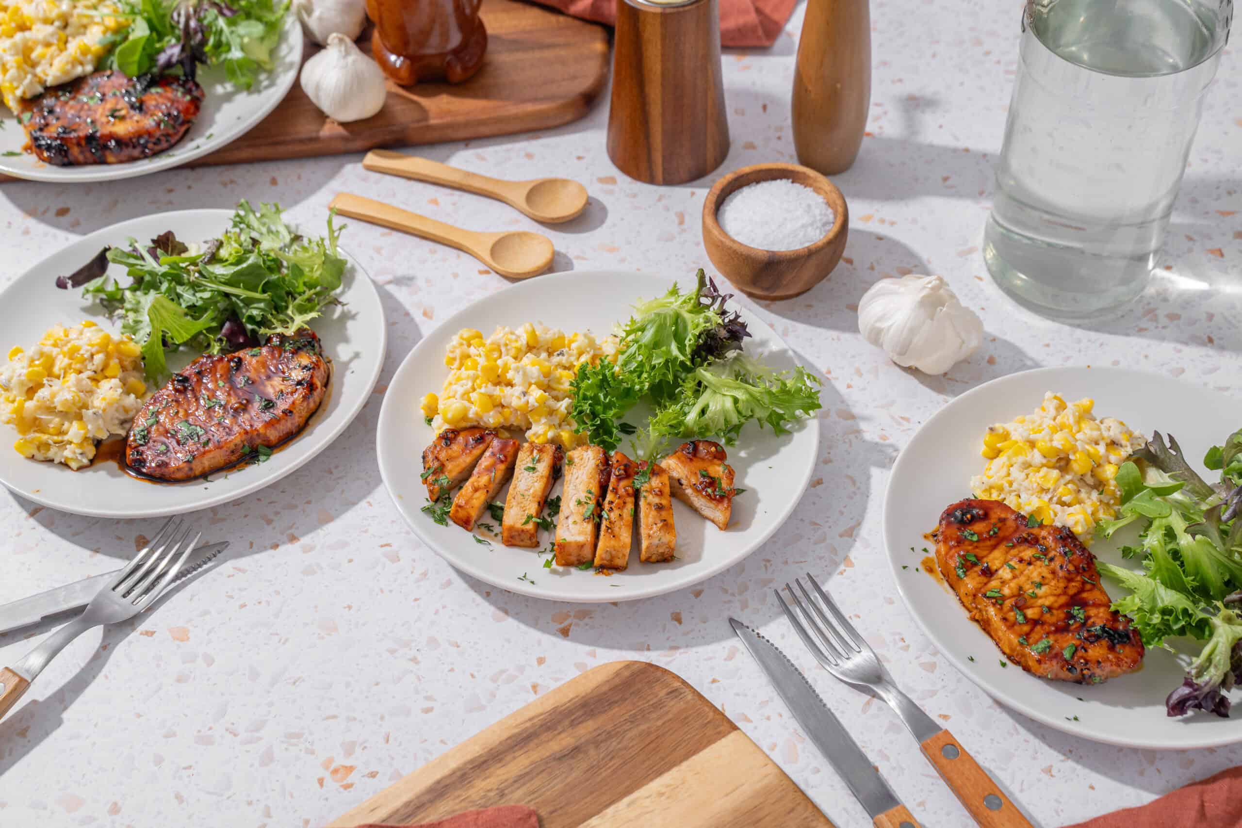 Grilled chicken with fresh salad, scrambled eggs, and herbs on white plates served at a bright tabletop with cooking ingredients and utensils.
