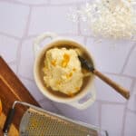 Creamy mashed potatoes with orange zest in a white ceramic bowl on a kitchen counter with fresh orange, grater, and other ingredients.