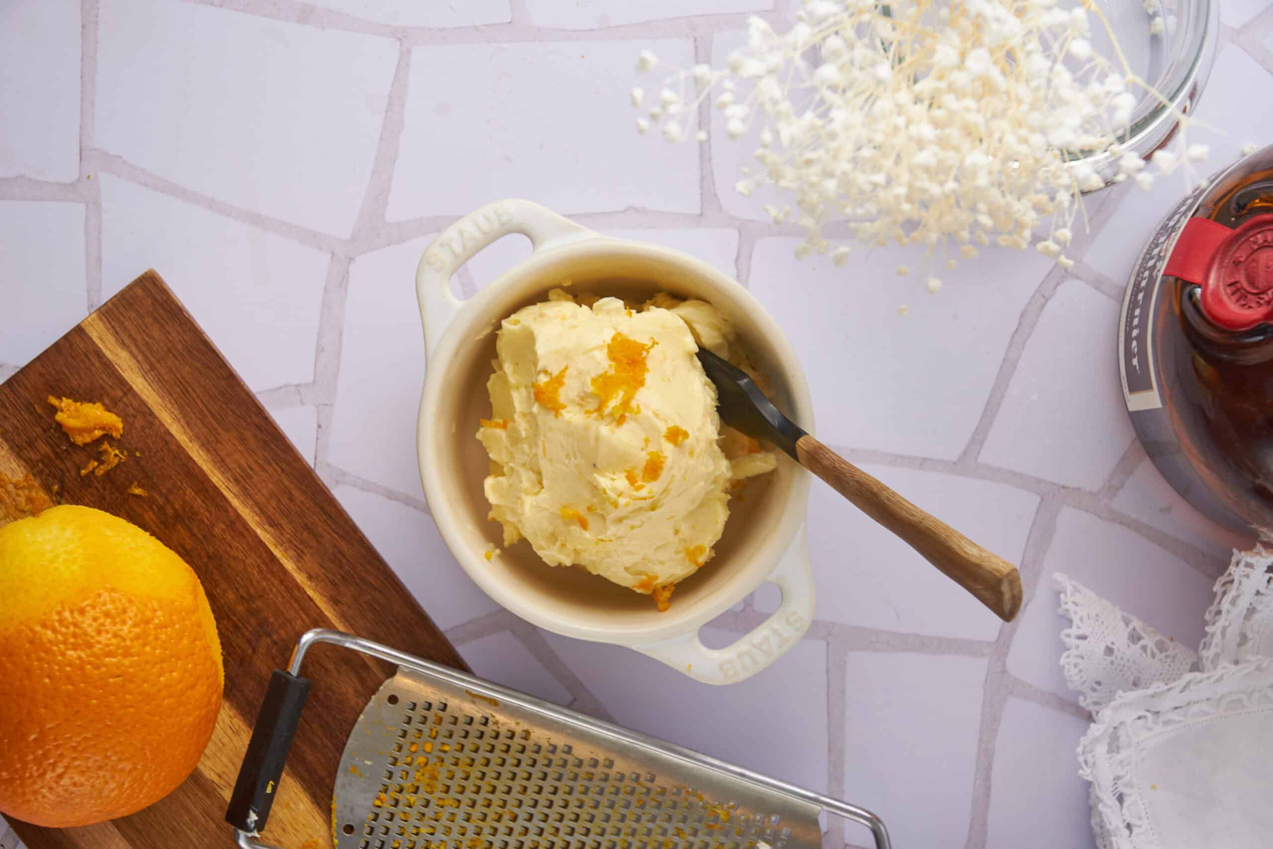 Creamy mashed potatoes with orange zest in a white ceramic bowl on a kitchen counter with fresh orange, grater, and other ingredients.