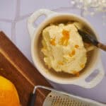 Creamy homemade orange-flavored ice cream in a small white ceramic bowl with a wooden-handled spoon, surrounded by a fresh orange, a wooden cutting board, and a metal grater, perfect for summer desserts.