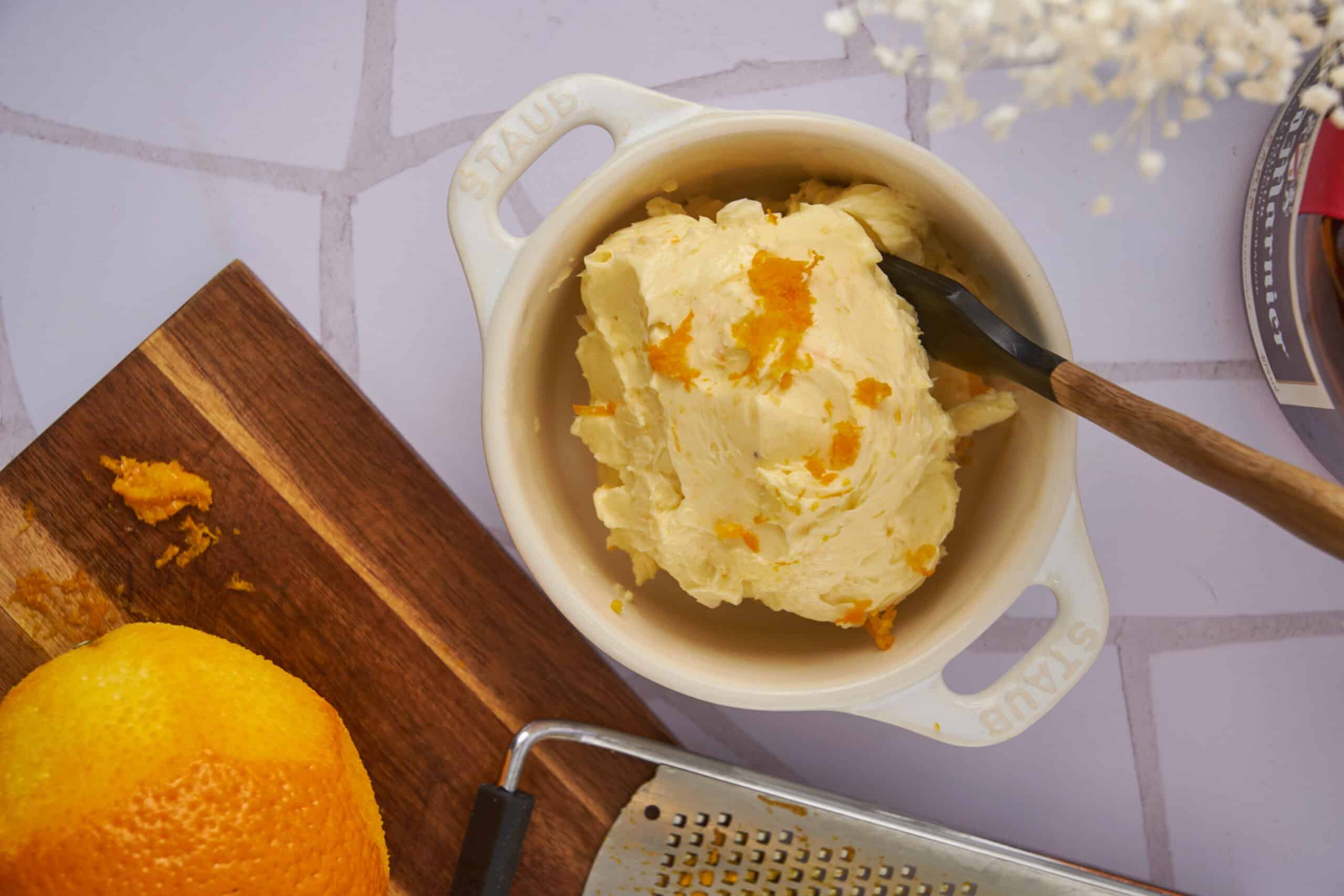 Creamy homemade orange-flavored ice cream in a small white ceramic bowl with a wooden-handled spoon, surrounded by a fresh orange, a wooden cutting board, and a metal grater, perfect for summer desserts.