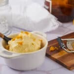Creamy homemade vanilla ice cream in a white bowl with a wooden-handled spoon, surrounded by a grater with orange zest, fresh pear, and baking ingredients on a white tiled surface.